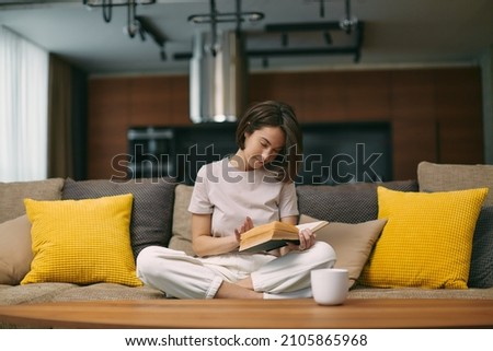 Modern young girl reading bestseller novel in paper book, enjoying weekend, relaxing on sofa at home. Self-education Royalty-Free Stock Photo #2105865968