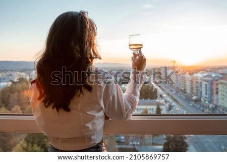 Brunette enjoying sunset. She is holding glass of wine next to window. Cityscape in the background.