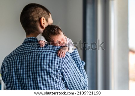 Back view of father holding his newborn baby while standing by the window in day at home - adult male holding sleepy infant child while looking trough the window waiting new life parenthood concept Royalty-Free Stock Photo #2105921150