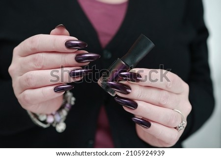 Female hand with long nails and dark purple burgundy manicure holds a bottle of nail polish
