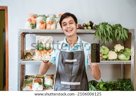 Male greengrocer showing vegetables and showing a phone screen