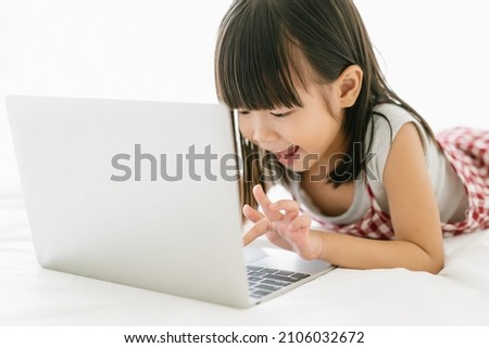 Portrait of little Asian girl enjoy playing game on notebook computer while lying on the bed in a relaxed atmosphere at home. Children Health care, kid learning activity, technology concept