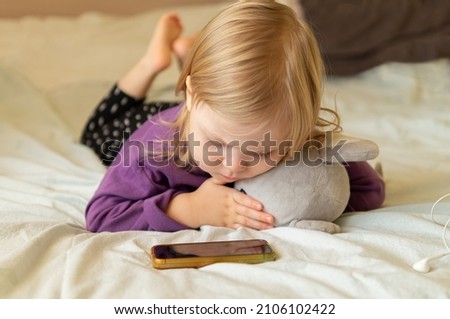 Beautiful little girl is lying in bed at home hugging a toy and using a mobile phone, taking selfies, watching cartoons, making a video call, browsing internet.