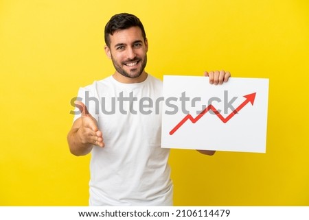 Young handsome caucasian man isolated on yellow background holding a sign with a growing statistics arrow symbol making a deal