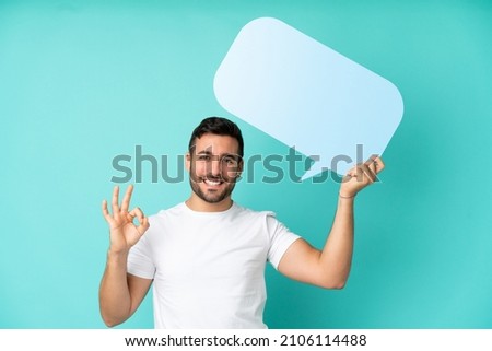 Young handsome caucasian man isolated on blue background holding an empty speech bubble and doing OK sign