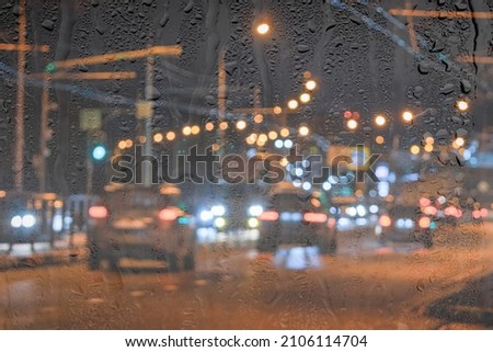 Blurry moving cars with headlights and lanterns reflecting on the wet asphalt in the night city behind rain-covered window. Rainy bad weather concept.