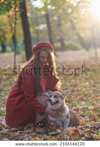 A girl with long red hair, in a red coat and beret, gently strokes her corgi dog in an autumn park. Golden autumn, walking the dog, friendship. beautiful autumn picture