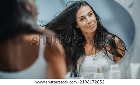 Beautiful Early Middle Aged Woman Looks into Bathroom Mirror Touches Her Lush Black Hair, Admires Her Looks. Concept for Happiness, Wellbeing, Natural Beauty, Organic Skin Care Products Royalty-Free Stock Photo #2106172052