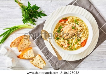 chicken noodle soup with carrots and scallion in a white bowl on a white wooden table with white bread and spoon, horizontal view from above, flat lay