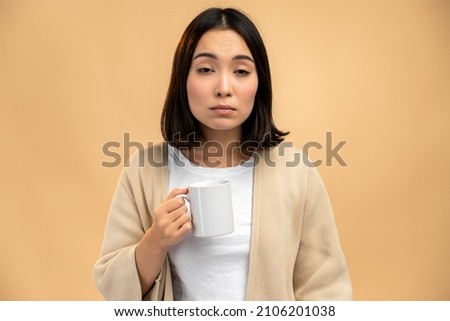 Sleepy asian ethnic female with sleepy eyes holding mug while drinking coffee early in morning against beige background  Royalty-Free Stock Photo #2106201038