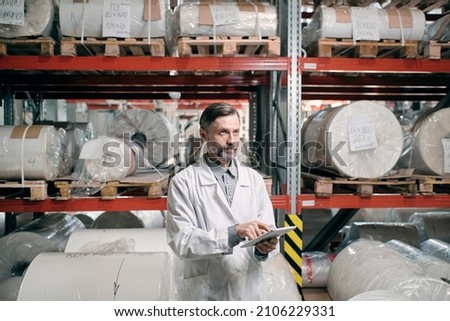 Portrait of male factory worker in white robe, using tablet for quality control and logistic purposes at polymer plastic manufacturing, standing between shelves with polymer rolls