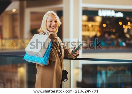 Making the retail connection. Young cheerful blonde shopper with paperbags scrolling in smartphone or looking through messages or adverts while shopping in the mall. 