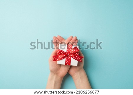 First person top view photo of valentine's day decorations young person's hands holding small white giftbox with red ribbon bow on crossed palms on isolated pastel blue background with copyspace