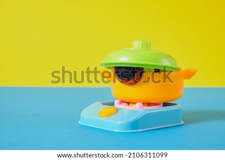 selective focus of friendly funny face concept, toy eyes in a plastic pot with a lid on the toy cooking stove, original idea minimal art copy space yellow and blue background