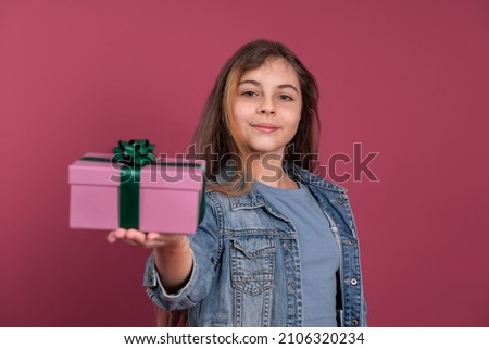 Cheerful 11 year old girl showing gift box to camera for concept of present for Mothers Day while looking at camera on pink background
