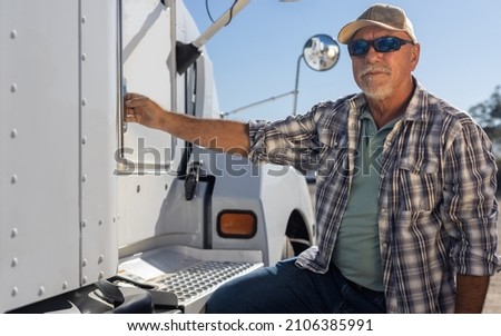 Hispanic middle aged semi truck driver sits next to truck. Experienced senior trucker next to big rig at the job. Portrait of transportation and logistics worker next to equipment on a sunny day. Royalty-Free Stock Photo #2106385991