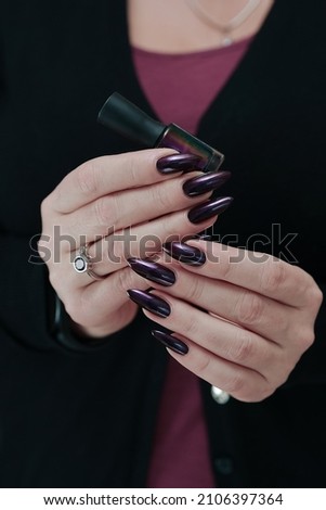 Female hand with long nails and dark purple burgundy manicure holds a bottle of nail polish