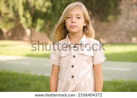 Joyful beautiful Caucasian little kid girl wearing dress standing outdoors  looking to the camera, thinking about something. Both arms down, neutral facial expression. Royalty-Free Stock Photo #2106404372