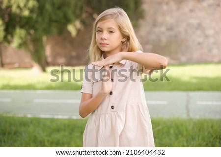 beautiful Caucasian little kid girl wearing dress standing outdoors  feels tired and bored, making a timeout gesture, needs to stop because of work stress, time concept.
