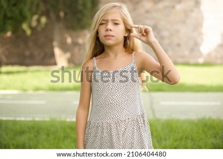 beautiful Caucasian little kid girl wearing dress standing outdoors  purses lip and gestures with hand, shows something very little.
