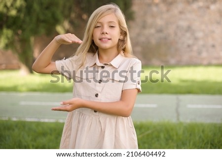 beautiful Caucasian little kid girl wearing dress standing outdoors  gesturing with hands showing big and large size sign, measure symbol.