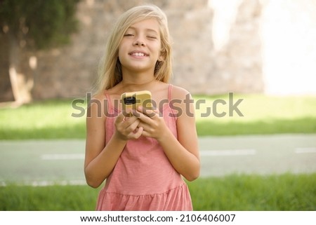 Pleased beautiful Caucasian little kid girl wearing dress standing outdoors using self phone and looking and winking at the camera. Flirt and coquettish concept.