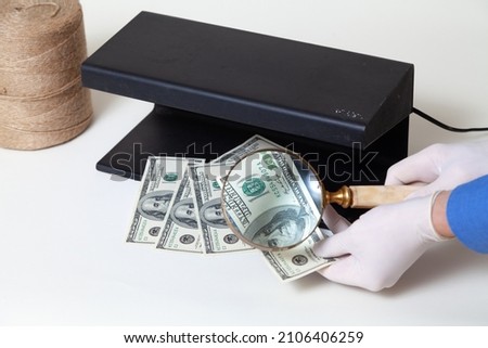 Currency authentication. Hands in rubber gloves hold a large magnifying glass on a bone handle over dollar bills. Near a metal detector.

