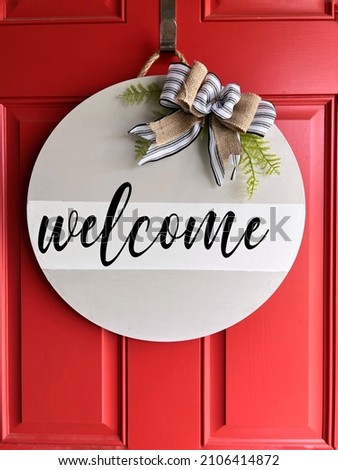 Homemade DIY craft round wood welcome sign with gray and white background and black cursive font reading "welcome" with a crafted farmhouse ribbon and green sprigs against a red door.