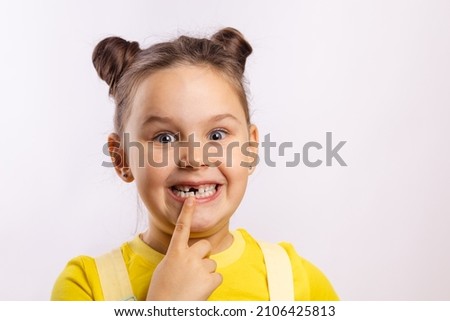 Female kid with opened mouth pointing at missing front baby tooth with finger smiling excitedly in yellow t-shirt on white background. First teeth changing. Going to dentist to do tooth treatment.  Royalty-Free Stock Photo #2106425813