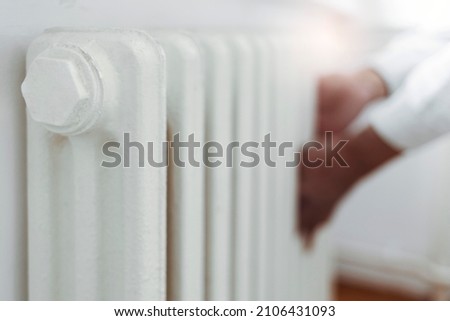 A man wearing a warm sweater warms cold hands on the heating radiator at home. Woman touches heating radiator indoors for checking the temperature. Selective focus. Heating system at home, concept.
