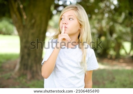 caucasian little kid girl wearing t-shirt standing outdoors silence gesture keeps index finger to lips makes hush sign. Asks not to share secret
