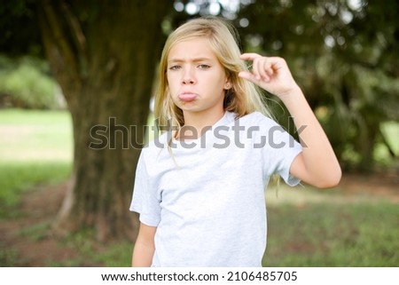 caucasian little kid girl wearing t-shirt standing outdoors purses lip and gestures with hand, shows something very little.
