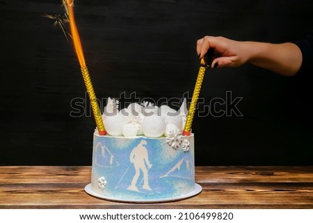 Female hand lighting firework candle, sparkler on fire in blue sponge vanilla creamy cake with round marshmallow, chocolate snowflakes, fir trees,mountain, skier picture,dessert on wooden table.
