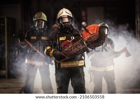 Group of professional firefighters wearing full equipment, oxygen masks, and emergency rescue tools, circular hydraulic and gas saw, axe, and sledge hammer. smoke and fire trucks in the background.  Royalty-Free Stock Photo #2106503558