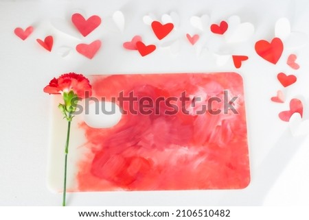 Valentines Day or wedding, invitation, scattered painted small cut out red, pink and white hearts with a red carnation on a colorful paint palette with copy space
