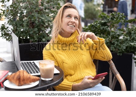 Happy girl sits in cafe with coffee and works on laptop. Dreaming blonde in yellow sweater. Thinking and smile face. Work online, freelancer style modern business woman. outdoor, outside