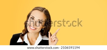Portrait of amazed smiling woman in black confident suit, pointing showing indicating copy space. Business concept. Orange yellow background. Brunette businesswoman. Executive employee office worker 