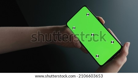 young woman sitting at the black table near window and holding smartphone with horizontal green screen