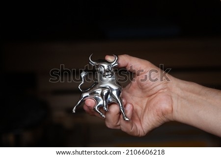 right hand holds a chrome bull figurine with horns and a raised tail on a black background

