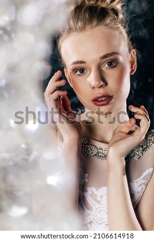 Light brown hair beautiful young girl in designer beige-white dress stands and sits on sofa and posing on the black wall background indoor  Royalty-Free Stock Photo #2106614918