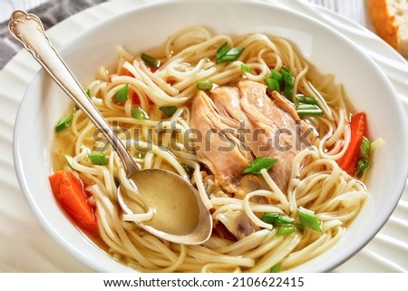 chicken noodle soup with carrots and scallion in a white bowl on a white wooden table with white bread and spoon, horizontal view from above, close-up