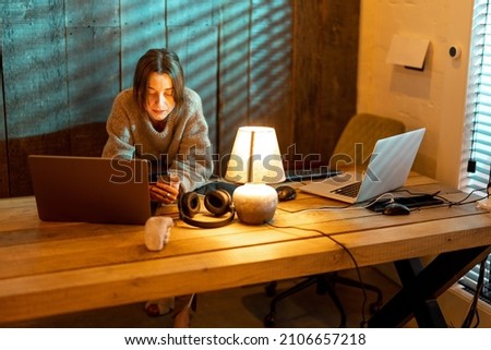 Young focused woman using smart phone while sitting at cozy home office during the nighttime. Concept of online work from home. Idea of cozy and style workplace Royalty-Free Stock Photo #2106657218