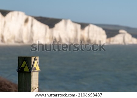 Yellow and black danger sign warning of erosion at the cliff edge. In the background Seven Sisters chalk cliffs at Seaford in East Sussex on the south coast of England UK. Photographed at Hope Gap.