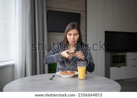 happy young woman sitting at home at the table having breakfast pancakes with orange juice. Takes a photo of your food with your smartphone. copy space