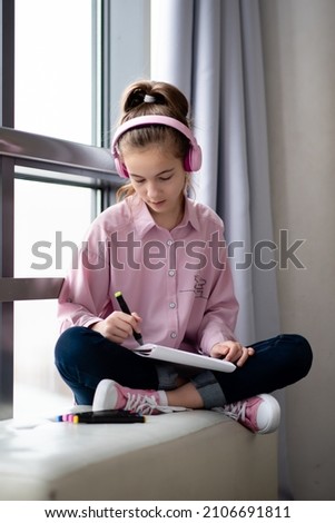 A cute teenage girl in a pink shirt and headphones sits near a window and draws with markers. Creation.