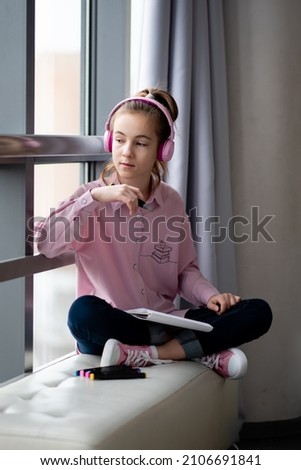 A cute teenage girl in a pink shirt and headphones sits near a window and draws with markers. Creation.