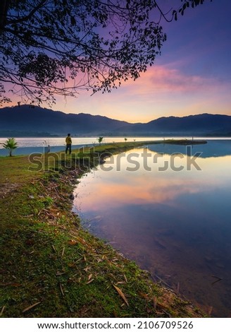 Lonely man standing in front of a lake and looking at the horizon. Alone man on the lake at sunrise looking a forest and mountains. Low light shooting. 