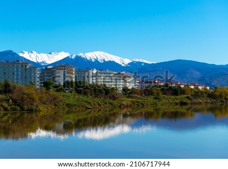 Beautiful daylight scenery of snowy Caucasus mountains with a wavy lake in front and modern buildings in the middle. Sochi, Adler Royalty-Free Stock Photo #2106717944