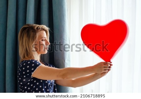 Young woman holding red heart shaped baloon against the window at home. Celebrating valentines day, wedding and love day, women's day, World Heart Day, birthday. Silhouetted against the sun indoor