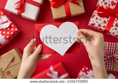 First person top view photo of saint valentine's day decor female hands holding red envelope and paper heart over present boxes on isolated red background with blank space